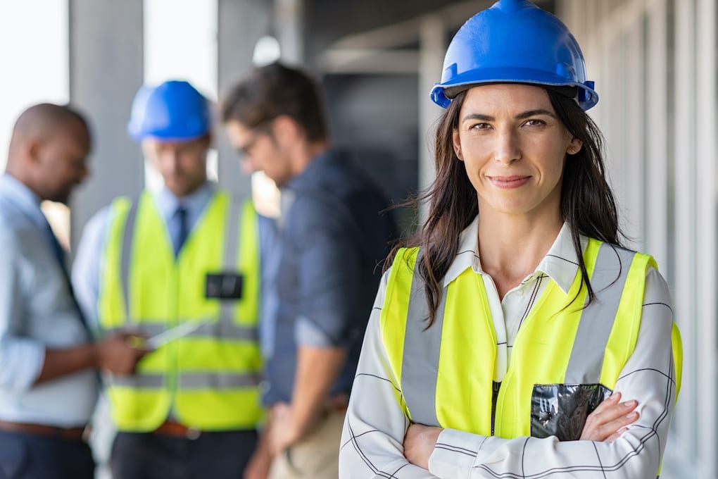 Portrait of woman engineer at building site looking at camera with copy space. Mature construction manager standing in yellow safety vest and blue hardhat with crossed arms; women in construction week