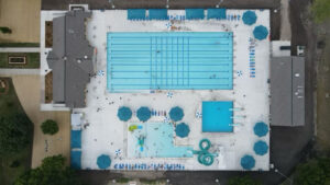 Drone footage of a commercial pool area, showing a large, well-maintained pool with clear, blue water surrounded by lounge chairs and umbrellas. The footage captures the pool's full layout, including surrounding landscaping, pathways, and nearby buildings, highlighting the space's design and functionality.