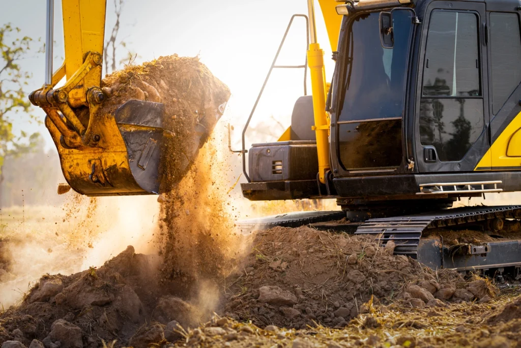 A yellow excavator operates in a field, its bucket dumping a load of soil, with dirt particles captured in mid-air against a sunlit background.