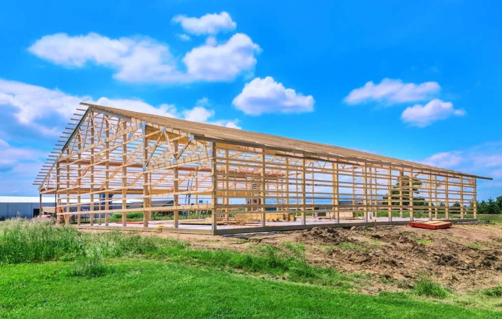 Large wooden frame structure under construction with clear blue sky and fluffy clouds overhead, situated on a grassy field with no people visible.