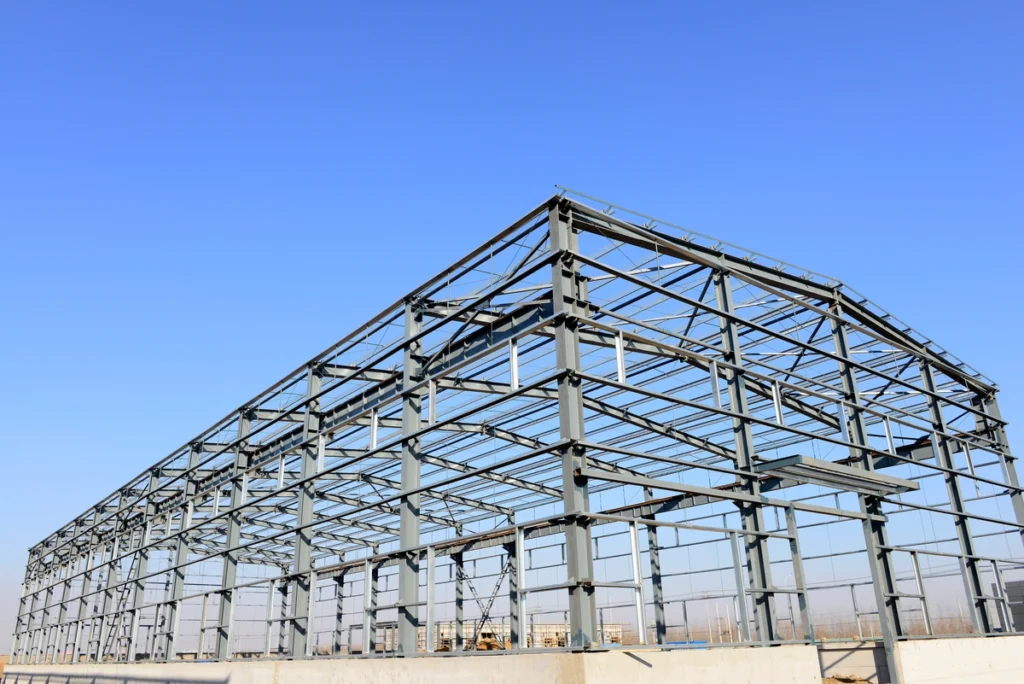 The image shows a large steel framework of a building under construction with a clear blue sky in the background, indicating an industrial or commercial project.