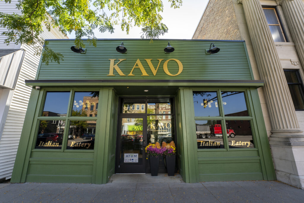Exterior view of KAVO Italian Eatery in St. Peter, Minnesota, featuring a green facade with large front windows, gold signage, and potted flowers by the entrance